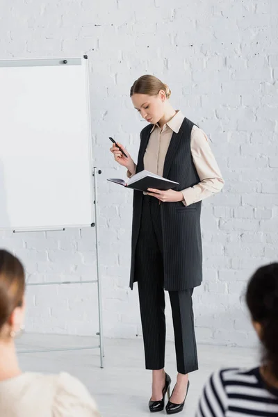 Young speaker looking at notebook while standing near flipchart and women on blurred foreground — Stock Photo