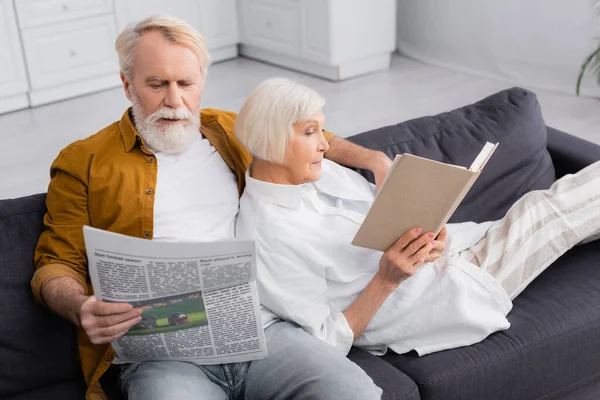 Homme âgé lisant le journal au premier plan flou près de la femme avec un livre sur le canapé — Photo de stock