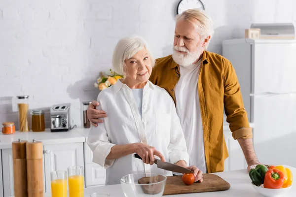 Anciano abrazando esposa cortando tomate mientras cocina en la cocina - foto de stock