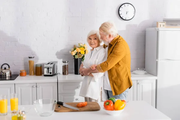 Uomo anziano che abbraccia sorridente moglie vicino tagliere e verdure fresche in primo piano sfocato — Foto stock