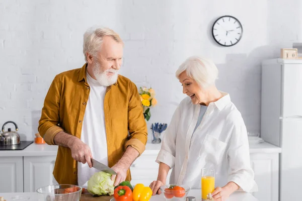 Homme coupant le chou près des légumes frais et femme souriante avec du jus d'orange dans la cuisine — Photo de stock