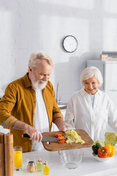 Senior man pouring vegetables in bowl while cooking near smiling wife in kitchen — Stock Photo