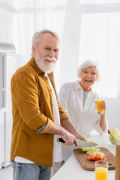 Älterer Mann blickt in die Kamera, während er Gemüse schneidet, neben lächelnder Frau mit einem Glas Orangensaft auf verschwommenem Hintergrund in der Küche — Stockfoto