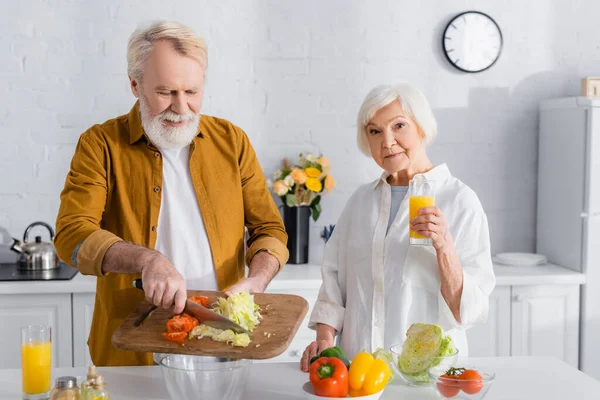 Senior woman holding glass of orange juice near husband pouring vegetables in bowl in kitchen — Stock Photo