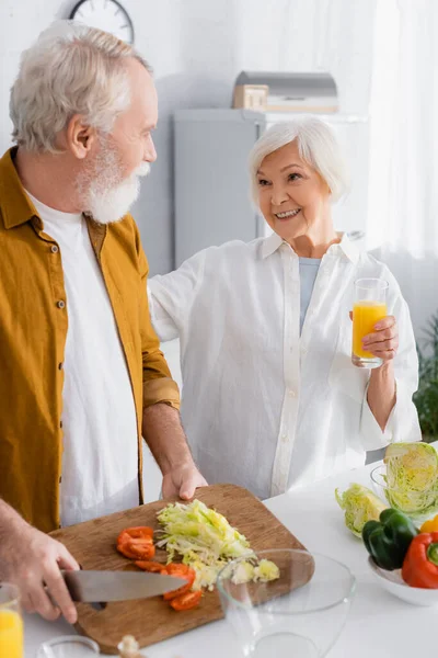 Smiling senior woman with orange juice looking at husband cutting vegetables in kitchen — Stock Photo
