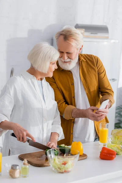 Hombre mayor sosteniendo teléfono inteligente cerca de esposa sonriente cocina en primer plano borrosa en la cocina - foto de stock