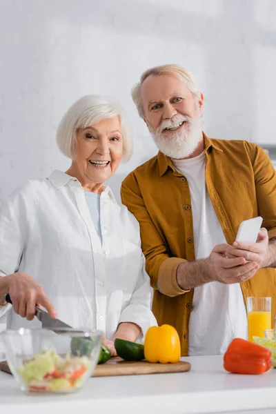 Улыбающаяся пожилая пара со смартфоном, смотрящая в камеру во время приготовления пищи на размытом переднем плане — стоковое фото