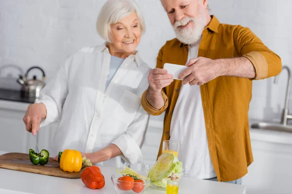 Fresh vegetables near smiling senior couple with smartphone cooking on blurred background — Stock Photo