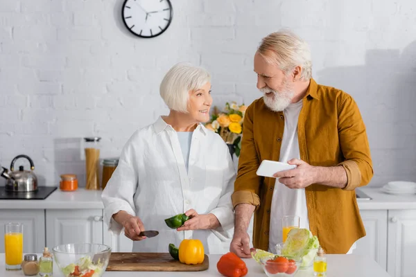 Hombre mayor sosteniendo teléfono inteligente cerca esposa sonriente con cuchillo y pimentón en la cocina - foto de stock