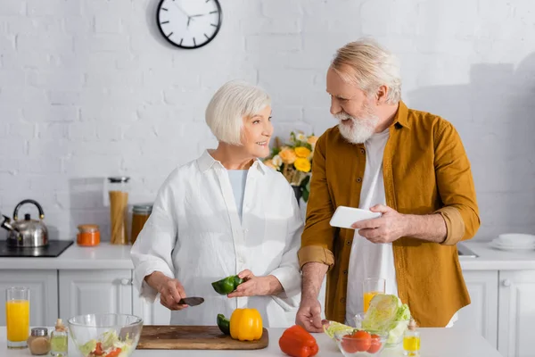 Senior man holding smartphone near smiling wife with knife and paprika in kitchen — Stock Photo