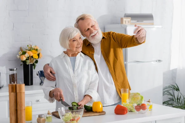 Senior man taking selfie on smartphone with smiling wife cooking in kitchen — Stock Photo