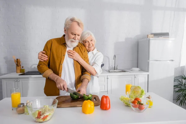 Donna anziana che abbraccia marito che cucina vicino al succo d'arancia in cucina — Foto stock