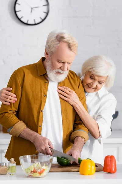 Старший мужчина режет паприку рядом с улыбающейся женой на кухне — стоковое фото