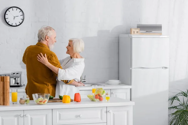 Pareja mayor abrazando cerca de verduras frescas y jugo de naranja en la cocina - foto de stock