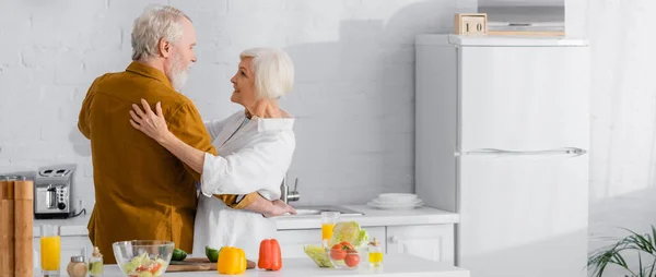 Позитивная пожилая пара, обнимающая рядом спелые овощи на кухонном столе, баннер — стоковое фото