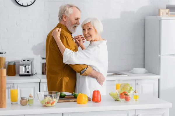 Senior woman smiling at camera while dancing with husband near vegetables in kitchen — Stock Photo