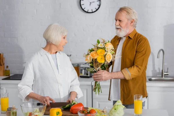 Старший мужчина преподносит цветы жене, режет овощи на кухне — стоковое фото