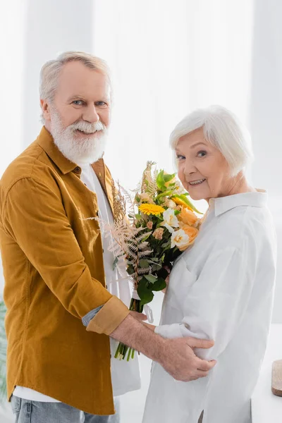 Couple aîné avec des fleurs souriant à la caméra dans la cuisine — Photo de stock