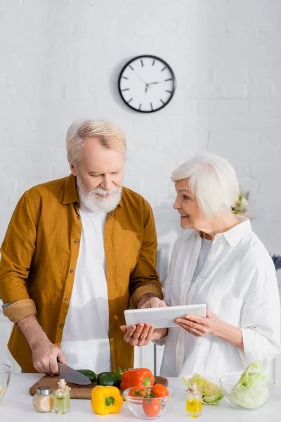 Lächelnde Seniorin hält digitales Tablet neben Ehemann beim Kochen in Küche — Stockfoto