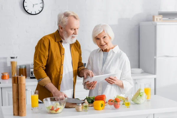 Mujer mayor con tableta digital de pie cerca de marido cocina en la cocina - foto de stock