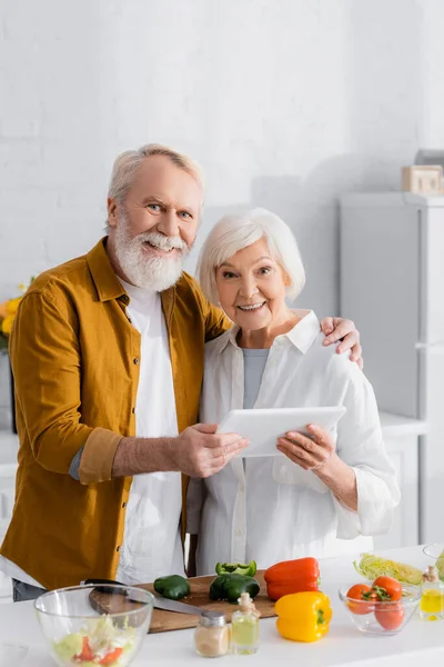 Fröhliches älteres Ehepaar hält digitales Tablet neben Schneidebrett und Gemüse auf verschwommenem Vordergrund in der Küche — Stockfoto