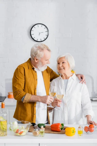 Старший мужчина обнимает жену во время звона с вином возле овощей на размытом переднем плане на кухне — стоковое фото