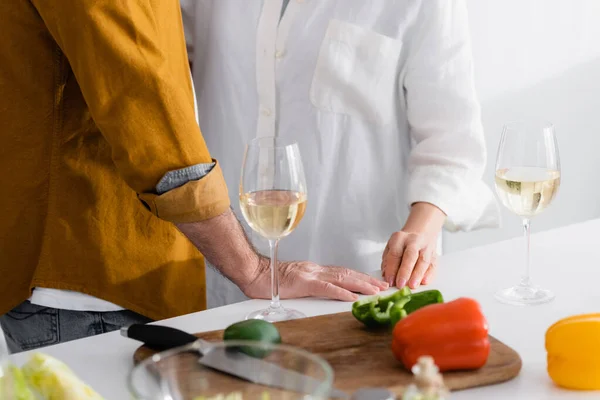 Vista cortada de casal sênior em pé perto de copos com vinho e páprica na mesa da cozinha — Fotografia de Stock