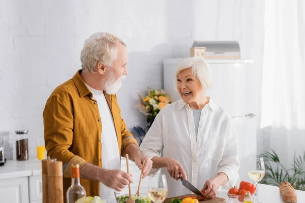 Веселая пожилая женщина режет овощи, а муж смешивает салат с вином на кухне. — стоковое фото
