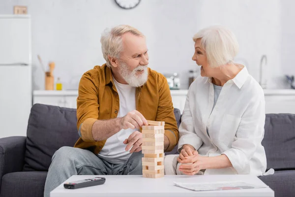 Allegro coppia di anziani giocare blocchi di legno gioco vicino telecomando e giornale in primo piano sfocato — Foto stock