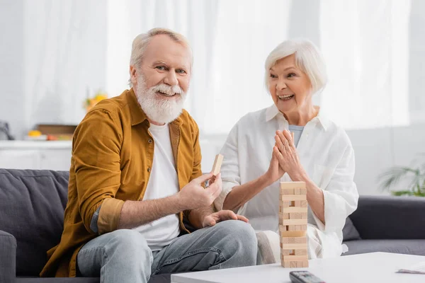 Senior man holding part of blocks wood game near cheerful wife in living room — Stock Photo