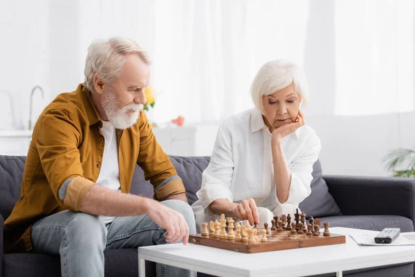 Pensive elderly couple playing chess near remote controller on blurred background — Stock Photo