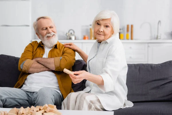 Senior woman holding part of blocks wood game near cheerful husband on couch — Stock Photo