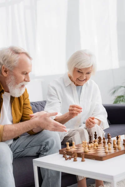 Femme âgée souriante assise près des échecs à bord et mari pointant la main sur le premier plan flou — Photo de stock