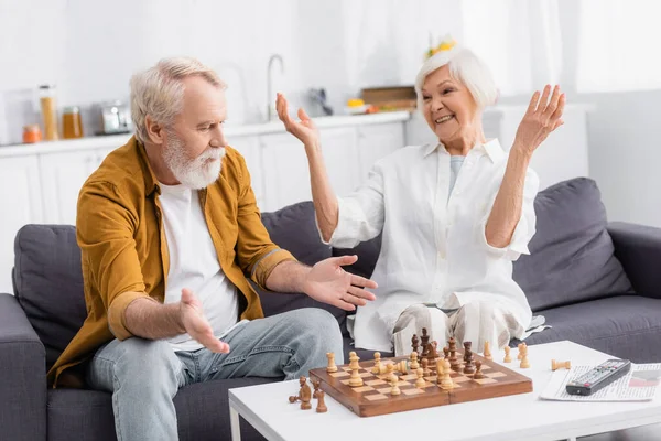 Senior homme pointant avec les mains vers les sangliers d'échecs près de femme souriante sur le canapé — Photo de stock