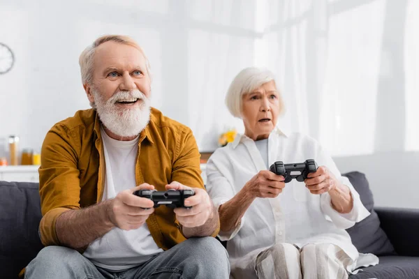 KYIV, UCRANIA - 17 DE DICIEMBRE DE 2020: Alegre hombre mayor jugando un videojuego cerca de su esposa molesta sobre un fondo borroso - foto de stock