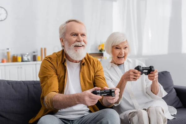 KYIV, UKRAINE - DECEMBER 17, 2020: Cheerful senior couple playing video game on sofa in living room — Stock Photo