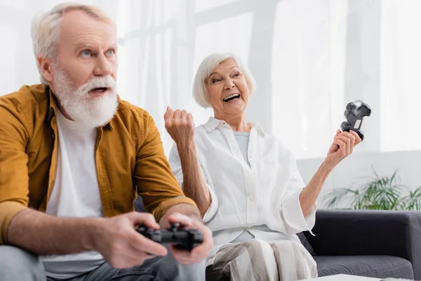 KYIV, UKRAINE - DECEMBER 17, 2020: Senior woman with gamepad showing yes gesture near husband on blurred foreground — Stock Photo