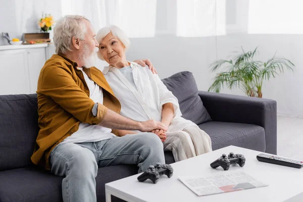 KYIV, UKRAINE - DECEMBER 17, 2020: Elderly couple hugging on couch near joysticks, remote controller and newspaper on coffee table — Stock Photo