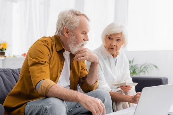 Umtriebiger Senior sitzt neben Ehefrau mit Taschenrechner und Laptop im verschwommenen Vordergrund — Stockfoto