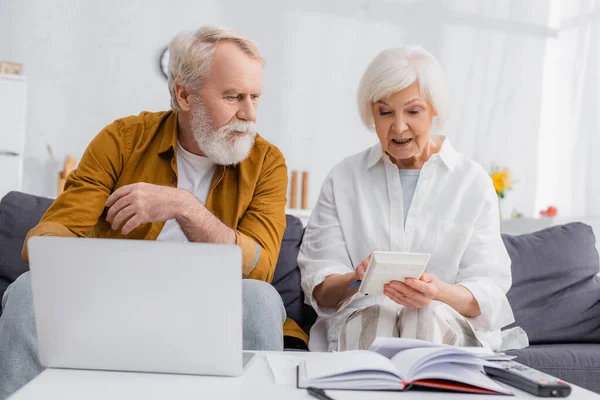 Femme âgée utilisant la calculatrice près de mari, ordinateur portable et ordinateur portable sur le premier plan flou — Photo de stock