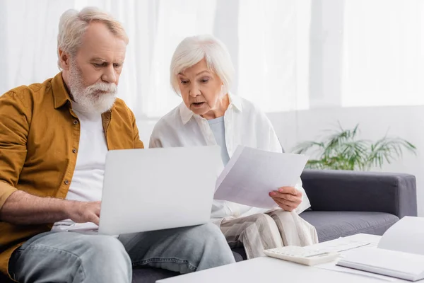 Hombre mayor usando portátil cerca de la esposa con papeles, calculadora y portátil en la mesa - foto de stock
