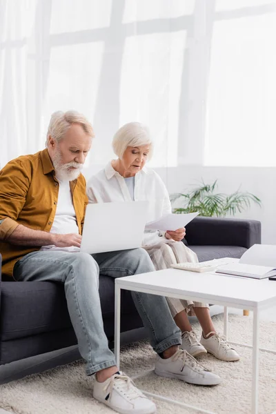 Elderly couple with laptop and papers sitting on couch — Stock Photo