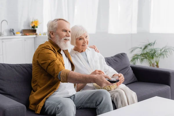 Smiling elderly couple watching tv with popcorn on couch — Stock Photo
