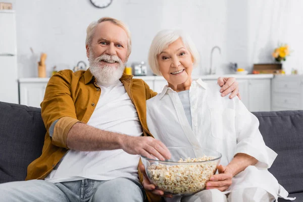 Alegre hombre mayor abrazando esposa con palomitas de maíz en el sofá - foto de stock
