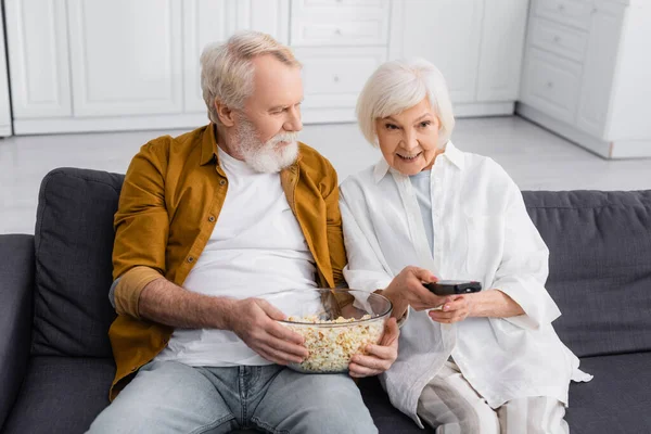 Smiling senior woman holding remote controller near husband with popcorn — Stock Photo