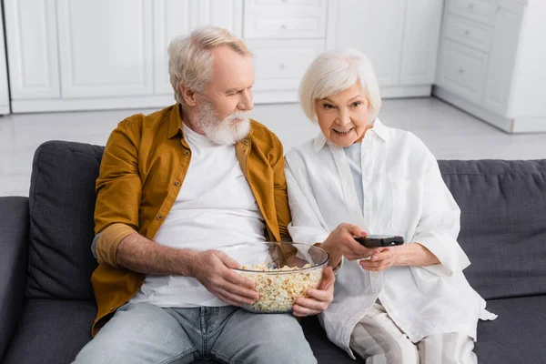 Усміхнена старша жінка тримає пульт дистанційного керування біля чоловіка з попкорном — стокове фото