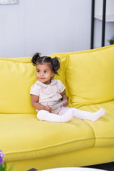 Felice ragazza afro-americana che tiene smartphone mentre siede sul divano — Foto stock