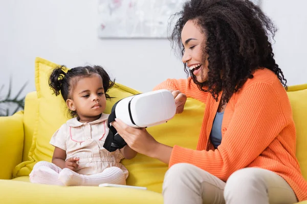 Cheerful african american mother holding vr headset near daughter in living room — Stock Photo