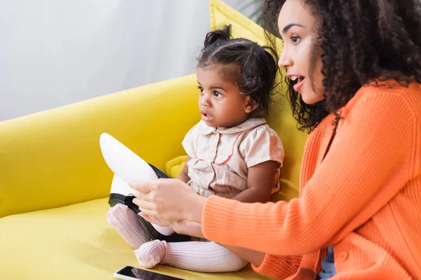 Erstaunt afrikanisch-amerikanische Mutter hält vr Headset in der Nähe Tochter im Wohnzimmer — Stockfoto