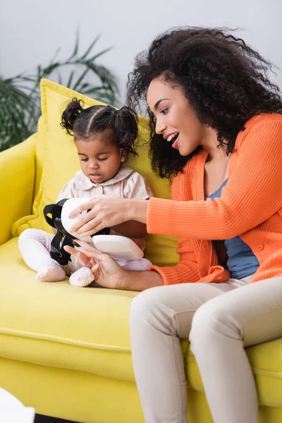 Asombrada madre afroamericana sosteniendo auriculares vr cerca de hija pequeña en sala de estar - foto de stock